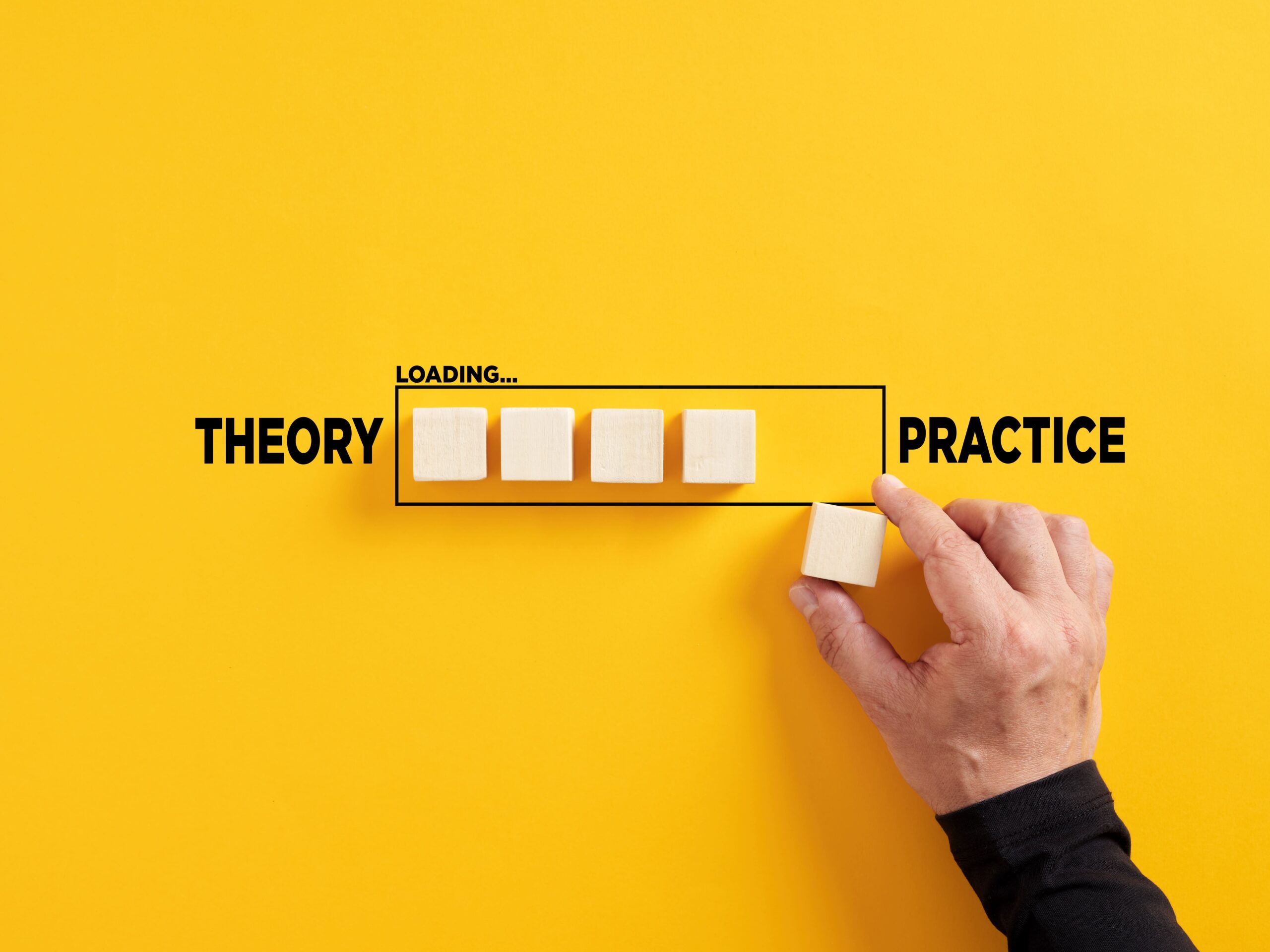 Business simulations – A hand places the last of four wooden blocks into a progress bar labeled "Loading...," with "Theory" on the left and "Practice" on the right, symbolizing the application of knowledge.